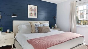 Un dormitorio con cama, mesitas de noche, una ventana con cortinas y un cuadro en la pared.
