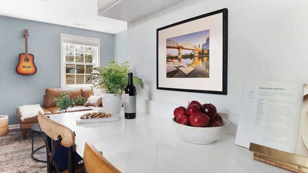 Una cocina moderna con comedor, que incluye una encimera blanca, una planta en maceta, una botella de vino y un bol de manzanas.