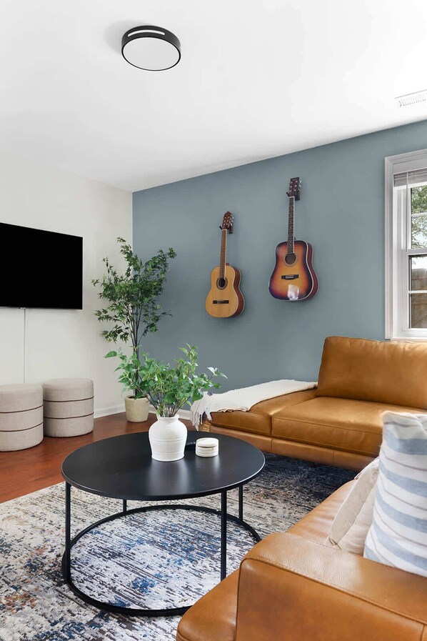 Una sala de estar moderna con un sofá de cuero marrón, una mesa de centro redonda negra, un televisor de pantalla plana grande y dos guitarras colgadas en la pared.