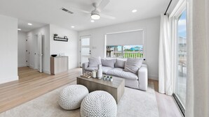 Un salon moderne avec un canapé gris, une table basse en bois et deux poufs ronds. On y trouve un ventilateur de plafond, un éclairage encastré et une grande fenêtre avec des rideaux.