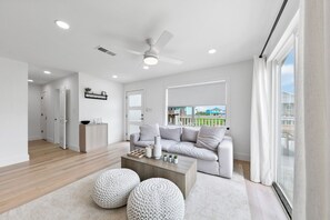 Un soggiorno moderno con un divano grigio, un tavolino in legno e due pouf rotondi. Si nota una ventola a soffitto, illuminazione a incasso e un'ampia finestra con tende.