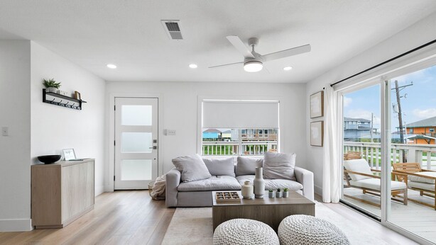 Un salon moderne avec un canapé gris, une table basse et deux poufs. On y trouve un ventilateur de plafond, une fenêtre donnant sur l’extérieur et une porte menant à une autre pièce.