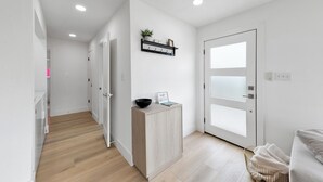 Un couloir moderne et bien éclairé, doté d’un plancher en bois, d’une porte blanche avec des panneaux en verre dépoli, d’une petite armoire en bois contenant un bol et une pancarte, et d’un panier en osier rempli de serviettes.
