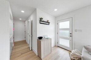 Un corridoio moderno e luminoso con pavimento in legno, una porta bianca con vetri satinati, un piccolo armadietto in legno con una ciotola e un cartello, e un cesto di vimini con asciugamani.