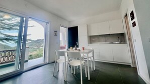 A modern kitchen with white cabinets, a dining area with a table and chairs, and a large sliding glass door leading to a balcony with a view of the hills. - Casa San Michele a Bagnaia (Portoferraio)