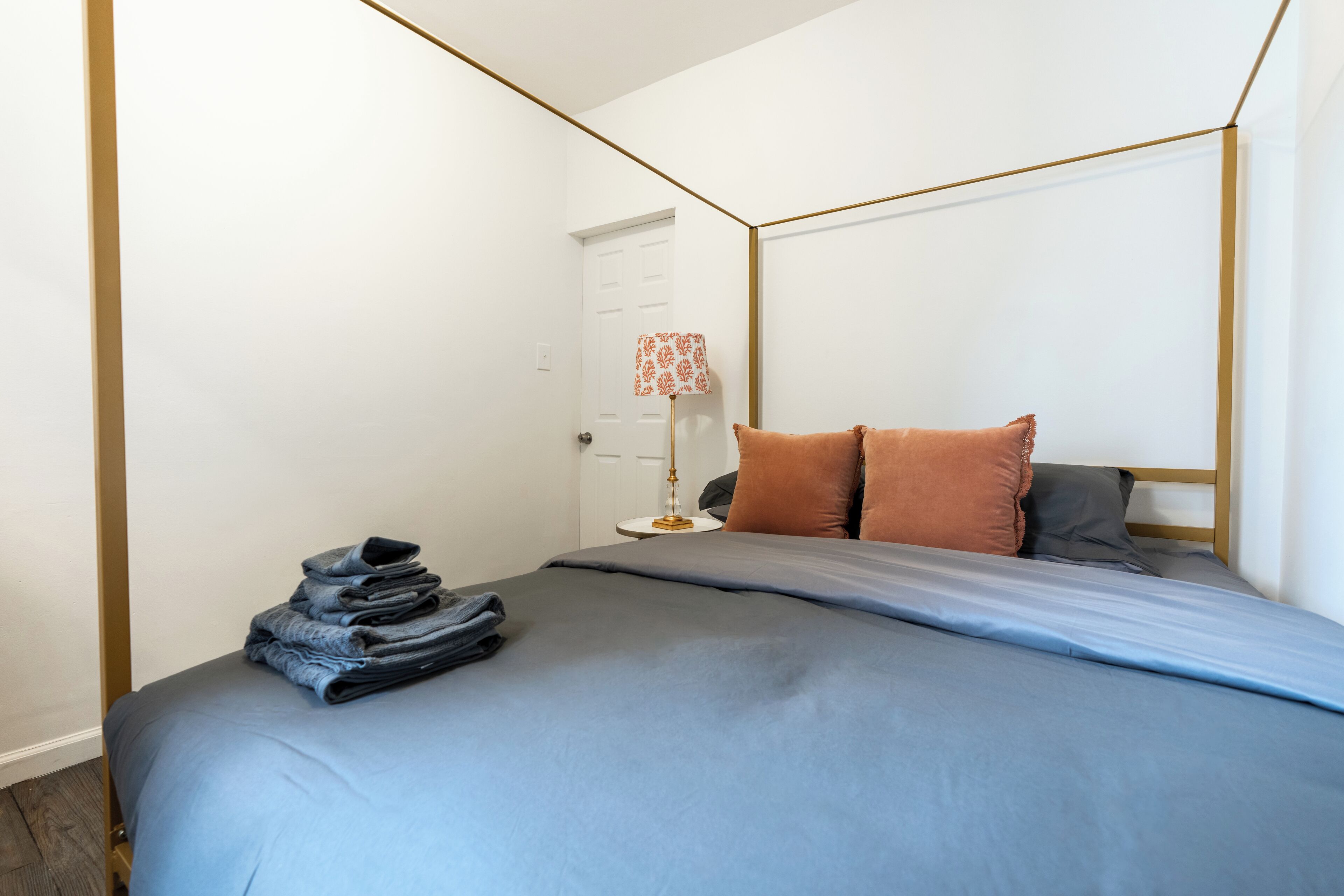 Una cama bien hecha con una colcha azul y almohadas, una lámpara con pantalla estampada, y una pila de ropa doblada a un lado.