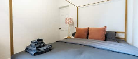 Una cama bien hecha con una colcha azul y almohadas, una lámpara con pantalla estampada, y una pila de ropa doblada a un lado.