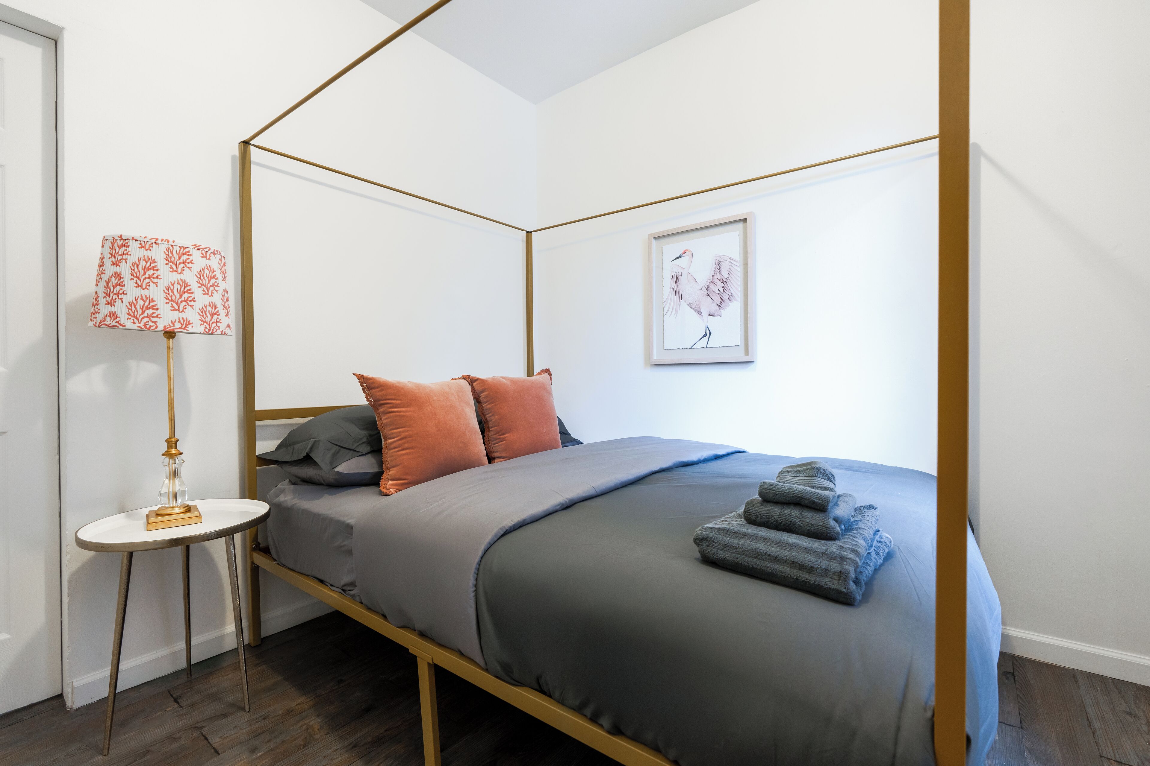 Una cama con dosel, ropa de cama gris, almohadas naranja y una obra de arte enmarcada en la pared.