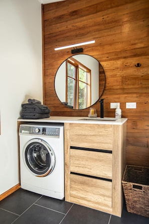 A compact laundry area with a front-loading washing machine, a wooden vanity with a mirror, and a small window. - The Piggery (Parapara)