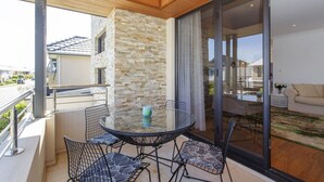 A balcony with a glass table and chairs, a stone wall, and a view of the street and neighboring houses.