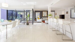 A modern, open-plan living and dining area with a large glass door leading to a balcony, white furniture, and a flat-screen TV mounted on the wall.