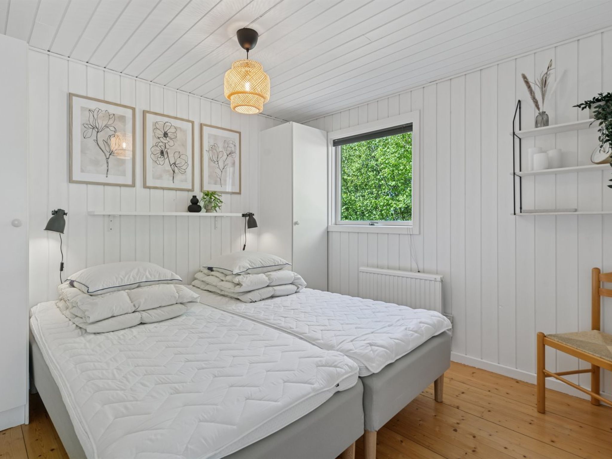 A bedroom with a double bed, white walls, wooden ceiling, and a window with a view of greenery.