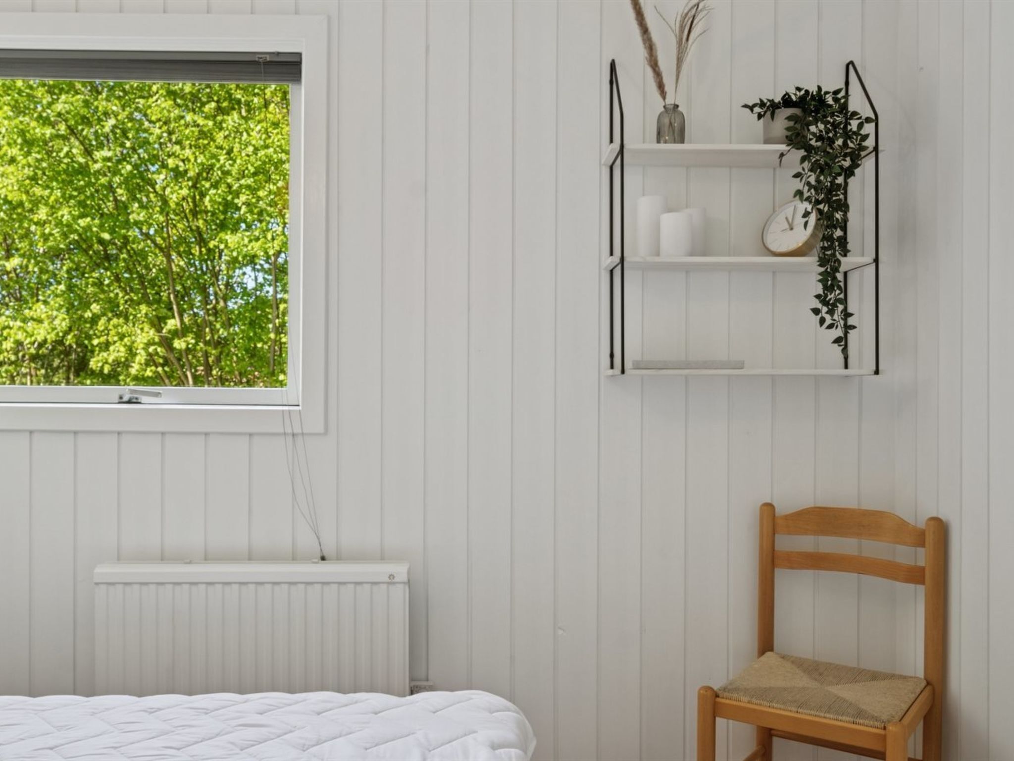 A bedroom with a bed, a radiator, a window with a view of trees, a wooden chair, and a wall-mounted shelf with plants and a clock.