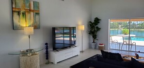 En moderne stue med et fladskærms-tv, et glasbord, en stor potteplante og udsigt til en swimmingpool.