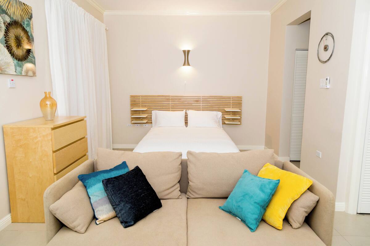 Habitación de hotel con cama, cómoda de madera, sofá color beige y cojines de colores, además de una lámpara fijada a la pared.