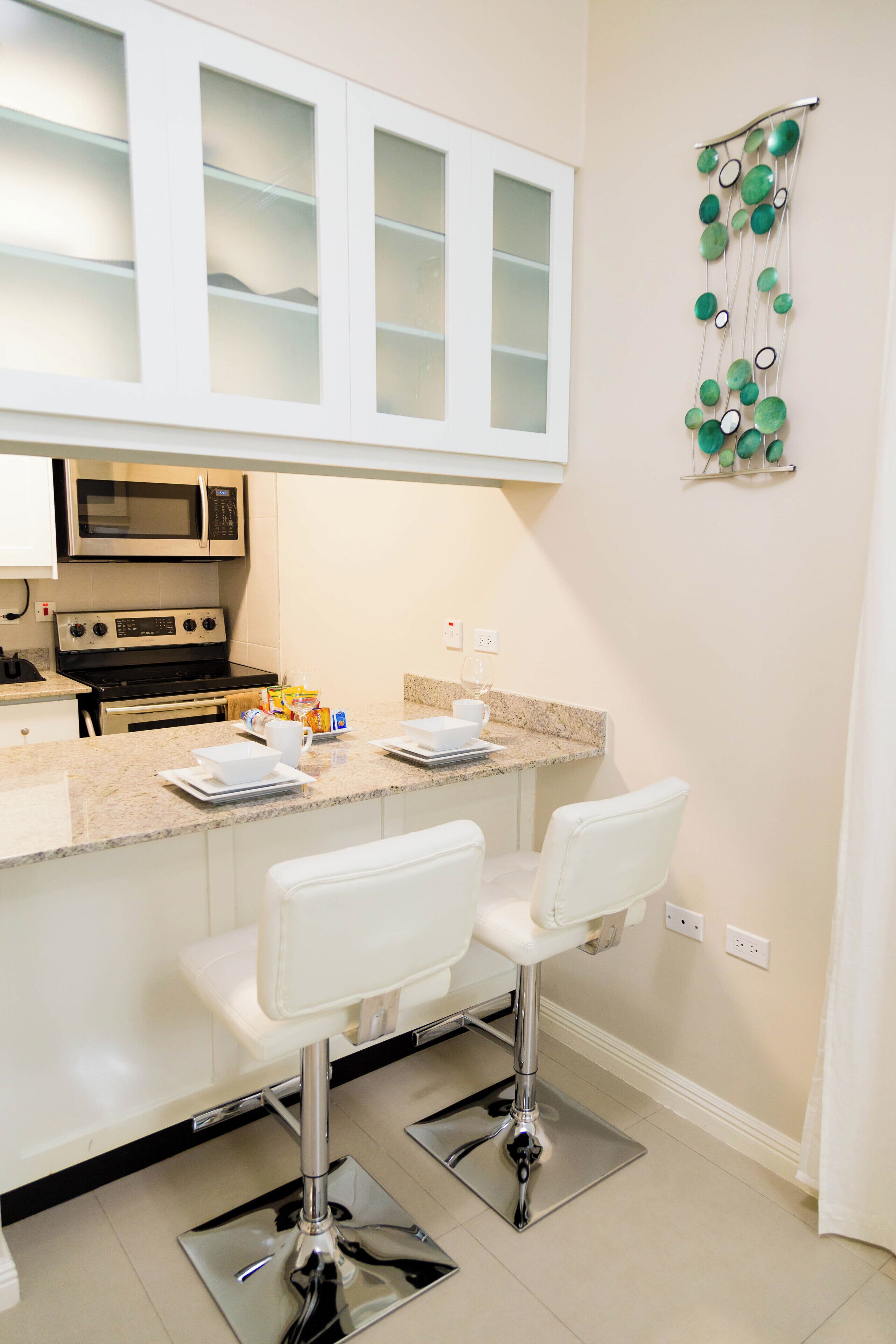 Una cocina moderna con armarios blancos, microondas integrado y encimera de granito. Hay dos taburetes blancos y una pieza decorativa en la pared.