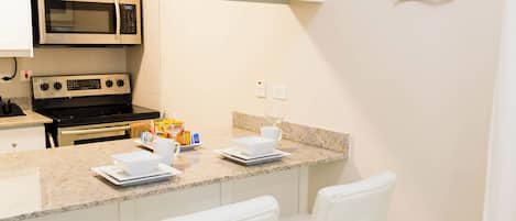 Una cocina moderna con armarios blancos, microondas integrado y encimera de granito. Hay dos taburetes blancos y una pieza decorativa en la pared.