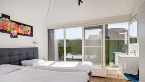A bedroom with a large bed, a desk, and a chair, a window with a view of a house, and a colorful abstract painting on the wall. - Kievit | Ruime bungalow met fijne tuin (Zwiggelte)