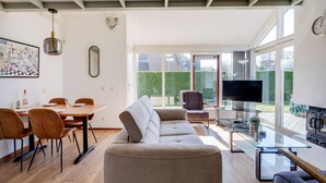 A modern living room with a sofa, a glass coffee table, and a TV. There is a dining area with chairs and a table, and a large window with a view of a garden. - Kievit | Ruime bungalow met fijne tuin (Zwiggelte)