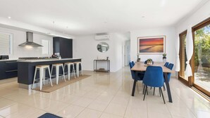 A modern kitchen with a central island, bar stools, and a dining area with blue chairs. - Wiltons Riverside family haven, pet friendly (Ocean Grove)