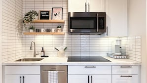 Une cuisine moderne avec des meubles blancs, un four à micro-ondes en acier inoxydable et une machine à café. Au-dessus de l’évier, on trouve deux étagères ornées d’objets décoratifs et d’une pancarte de bienvenue.