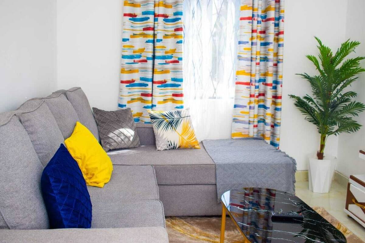 Ein modernes Wohnzimmer mit einem grauen Sofa, bunten Vorhängen, einem kleinen Tisch und einer Topfpflanze.