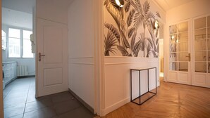 Un couloir avec une porte blanche, un mur orné d’une fresque représentant des feuilles tropicales, une petite console noire et un parquet en bois disposé en chevrons.