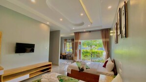 A hotel room with a TV, a wooden bench, two beds with pillows, a green rug, and a balcony with a view of greenery. - Finance Boutique (Kampot)