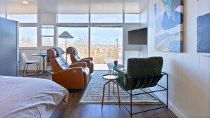 A modern hotel room with a large bed, a massage chair, a green armchair, and a small table.