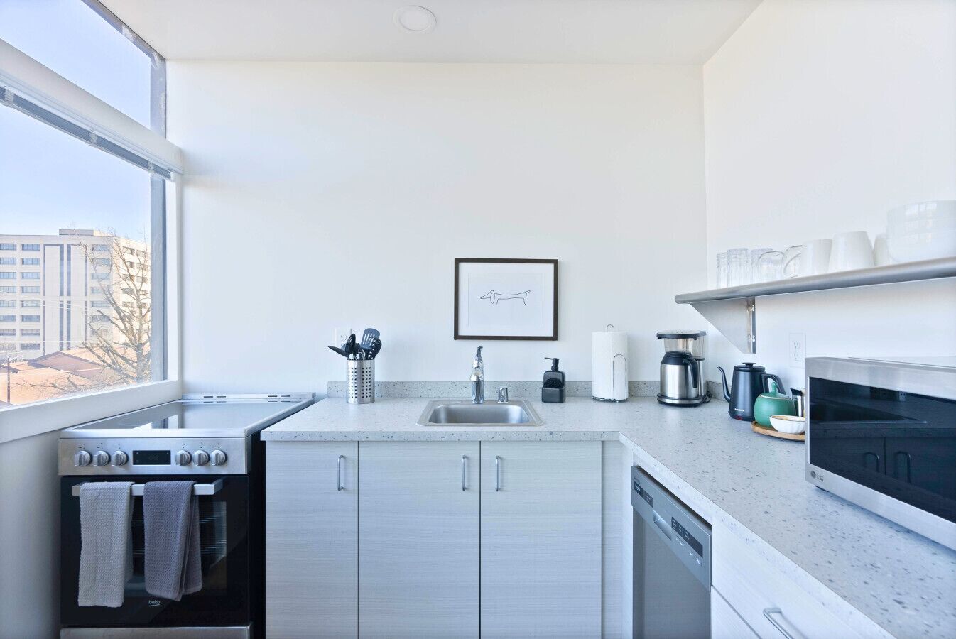 A modern kitchen with a stainless steel stove, a sink, a dishwasher, and a microwave. There is a window providing a view of buildings outside.