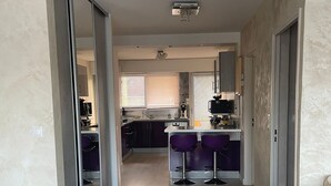 Una cucina moderna con isola centrale, zona pranzo con sedie viola e un corridoio che conduce a una camera da letto.