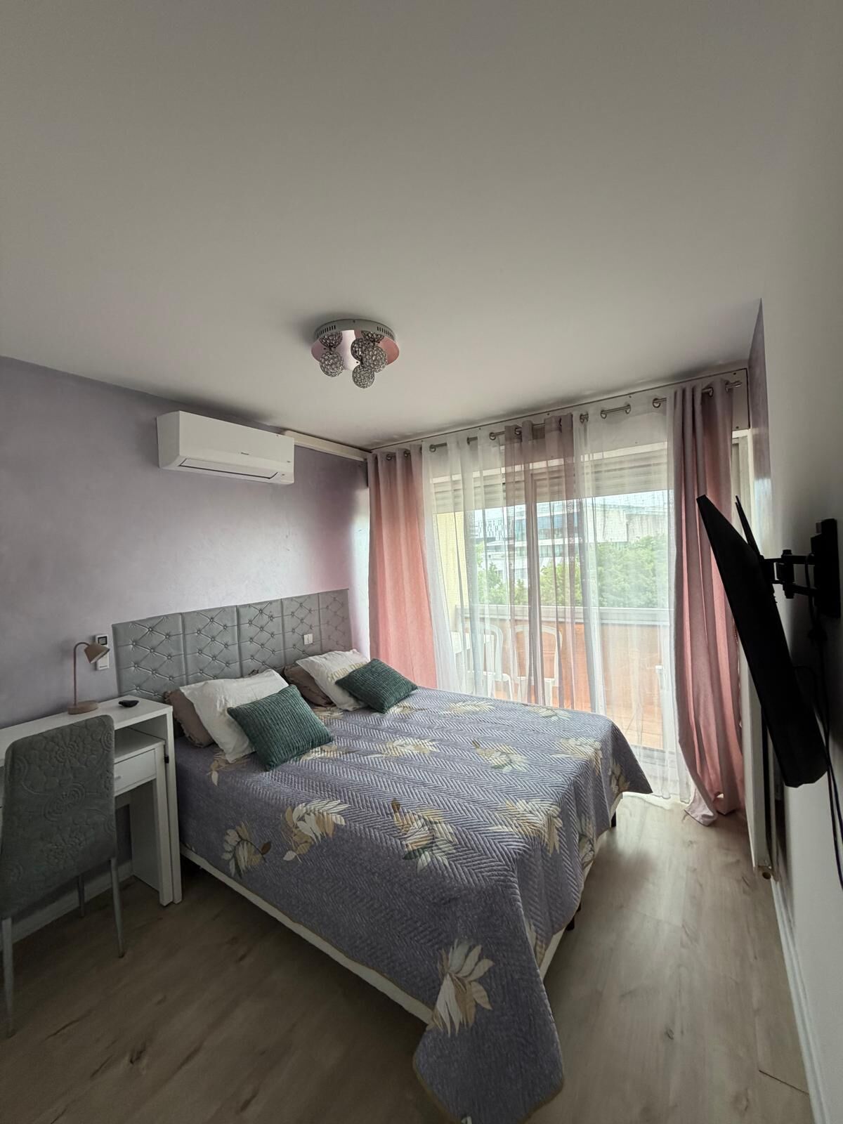 Quarto com cama, escrivaninha, cadeira, televisão e janela com cortinas.