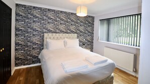 Ein ordentlich bezogenes Bett mit weißen Leinen und Handtüchern, ein gepolstertes Kopfstück, eine Wand mit Ziegeloptik, ein Fenster mit Jalousien und eine Hängeleuchte.