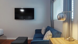 Uma sala de estar moderna com televisão de tela plana na parede, um sofá azul com almofadas estampadas, um pequeno banquinho e uma mesinha de madeira com abajur.