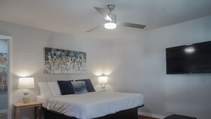 Quarto com cama, ventilador de teto, televisão fixada na parede e um quadro.