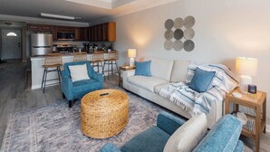 Un salon moderne avec un canapé, des fauteuils et une table basse. On aperçoit une cuisine à l’arrière-plan, avec des placards et des appareils électroménagers.