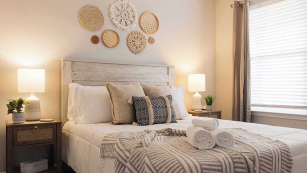 Un lit bien fait, avec une couverture à rayures et des serviettes pliées, deux tables de chevet avec des lampes et de petites plantes, et des œuvres d’art décoratives accrochées au mur au-dessus du lit.