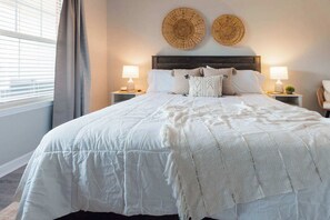 Un letto rifatto con un copriletto bianco e cuscini, due decorazioni murali tessute sopra e una finestra con tende a sinistra.
