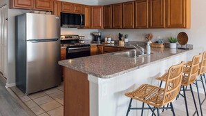 Une cuisine moderne avec des meubles en bois, un réfrigérateur en acier inoxydable, un plan de travail en granit et un coin bar avec des chaises en osier.