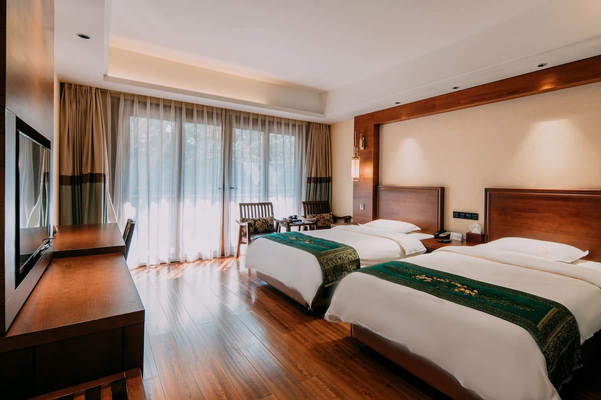 A hotel room with two beds, wooden flooring, a desk, and a large window with curtains.