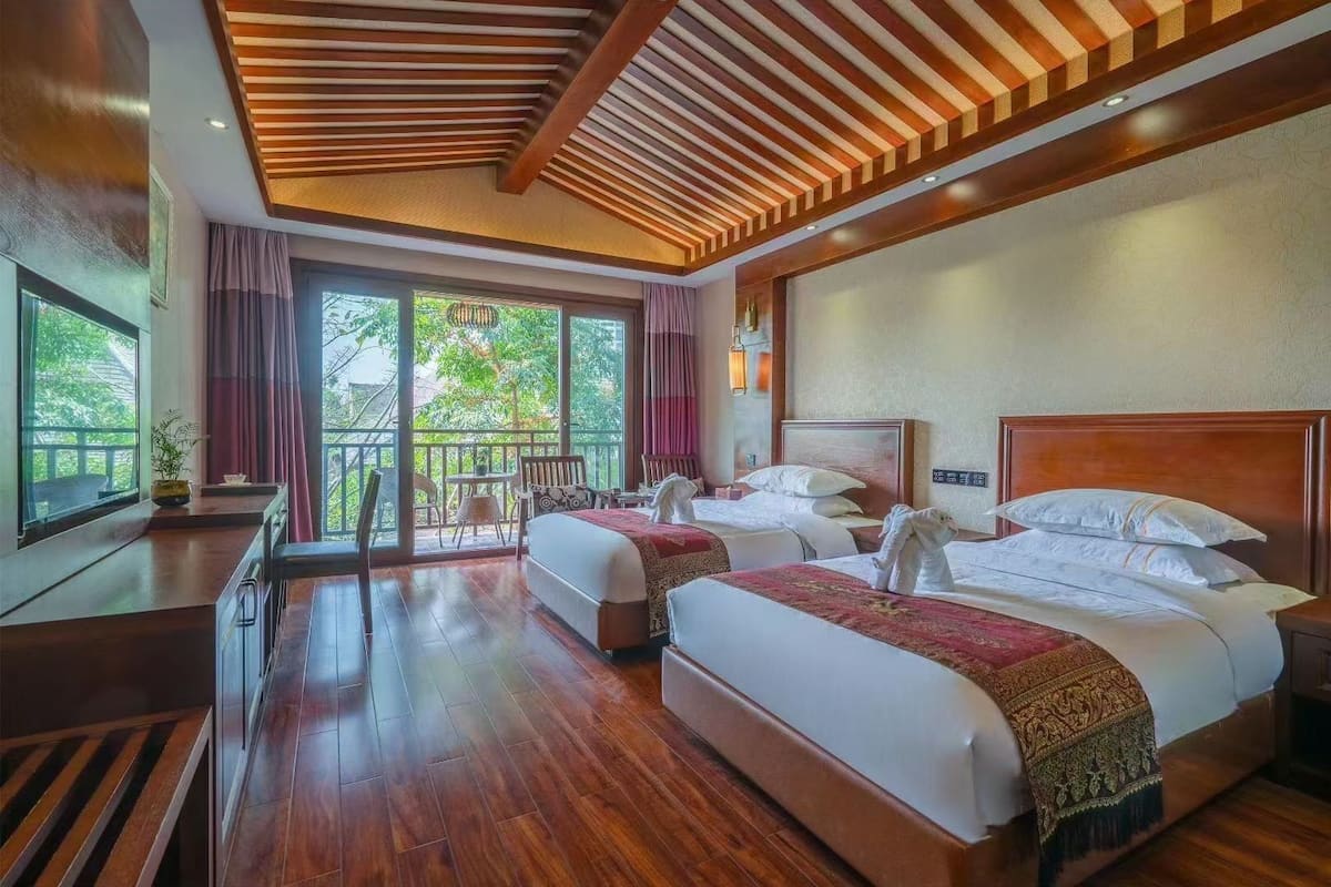 A hotel room with two beds, a desk, a chair, and a balcony with a view of greenery.