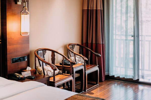 A hotel room with a wooden floor, a bed, two wooden chairs, a small table with a tissue box and remote controls, a wall-mounted light fixture, and a window with sheer curtains. - XiShuangBanNa BaoYu Vaction Hotel (Xishuangbanna Dai)