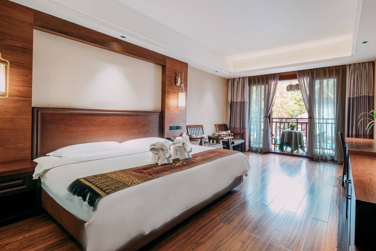 A modern hotel room with a large bed, wooden flooring, a desk, and a balcony with curtains.