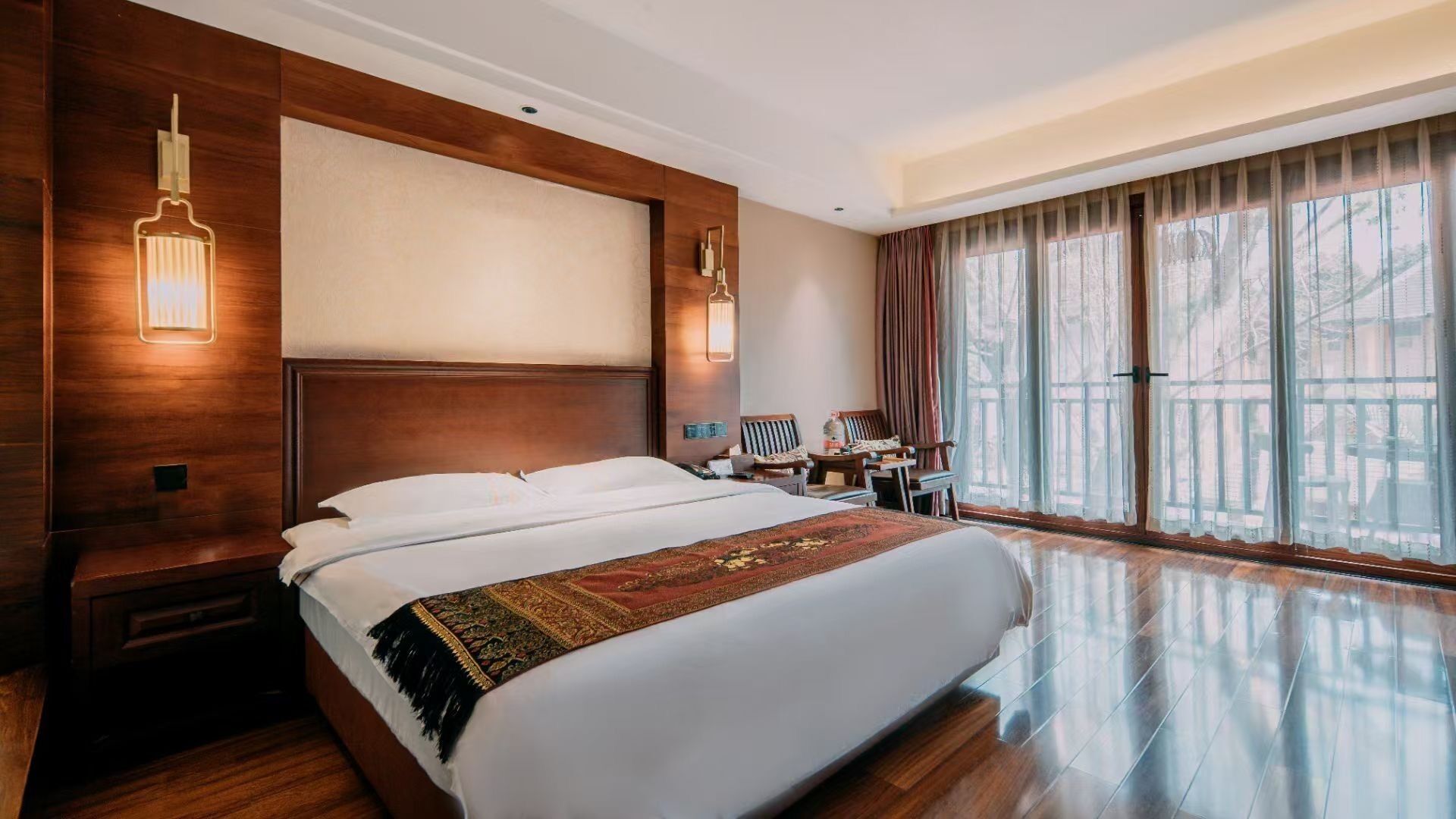 Une chambre d’hôtel avec un grand lit, une tête de lit en bois, deux lampes de chevet, un bureau avec une chaise et une grande fenêtre avec des rideaux.