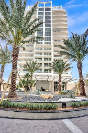 A multi-story hotel with a modern design, featuring balconies and a landscaped garden with palm trees.