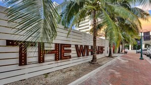 A sign with illuminated letters spelling "THE WHARF" against a white wall, with palm trees and a brick pathway in the foreground. - Steps to Beach Studio Suite | Pool & Las Olas (Fort Lauderdale)