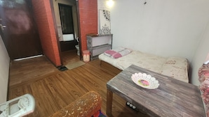 Un espacio habitable pequeño con piso de madera, un sofá tapizado con motivos florales, una mesita con un mantel de encaje, un banco y una puerta que da a un baño.