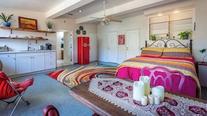 Un dormitorio con una cama grande, una mesa de madera con velas, una silla roja, una kitchenette y un ventilador de techo.