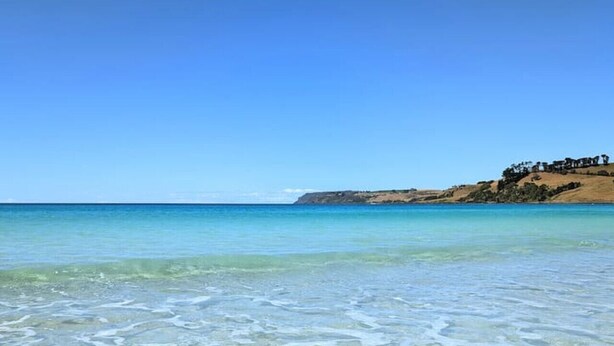 Uma praia com águas cristalinas e rasas, e uma colina distante coberta de árvores.