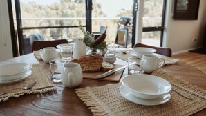 Mesa de jantar posta com pratos brancos, xícaras e um pão, com vista para árvores através de grandes janelas.